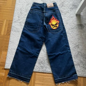 Mörkblå jeans med eldpatch från Bad Boy - Coola mörkblå jeans från Bad Boy med en unik eldpatch på bakfickan. De är nya och oanvända men prislappen är tyvärr borttagen🥲Byxorna har en loose fit och fransade kanter vid bensluten. Perfekta för en avslappnad stil med en edgy touch. 