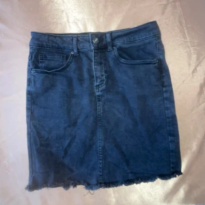 Svart jeanskjol - Svart  jeanskjol med rå kant och klassisk femficksdesign. Perfekt för en avslappnad stil. Kjolen har en knapp och dragkedja framtill.