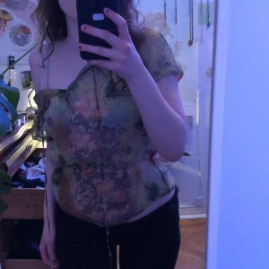 Blommig blus - Säljer en blommig offshoulder blus med korta ärmar och knytband. Blusen har ett färgglatt mönster i grönt, rosa och orange på en transparent bas. Perfekt för en somrig look!