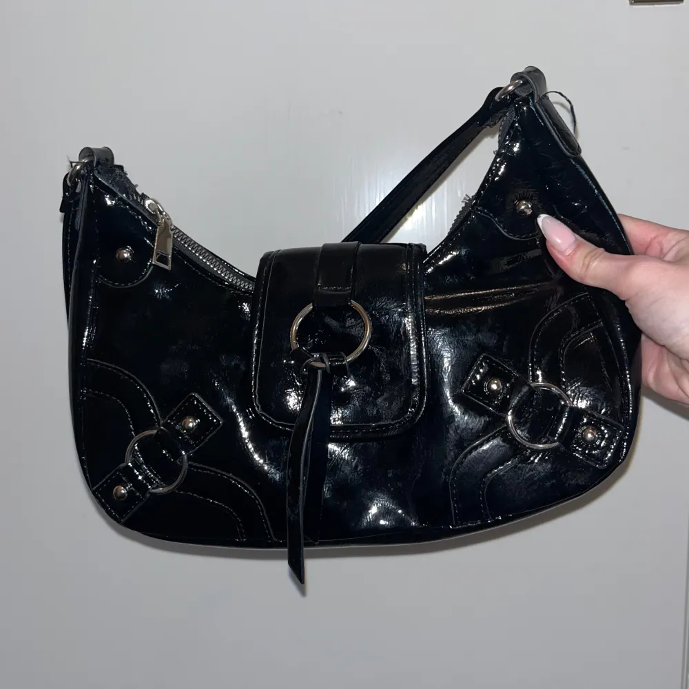 Snygg svart axelväska i glansigt material med dragkedja och dekorativa ringdetaljer. Perfekt för att ge en stilren touch till din outfit. Använd fåtal gånger. . Laukut & Käsilaukut.