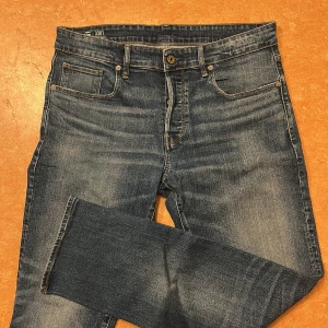 Blå jeans från G-Star RAW - Snygga blå jeans från G-Star RAW, modell 3301 Straight. Klassisk femficksdesign med knappgylf och läderpatch med logga baktill. Perfekta för en avslappnad stil.