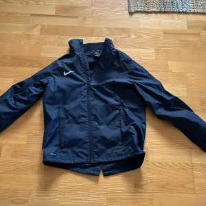 Mörkblå vindjacka från Nike - Säljer en mörkblå vind/regnjacka från Nike med dragkedja och hög krage. Har en avtagbar luva. Jackan har långa ärmar och är tillverkad i ett lätt syntetmaterial. Perfekt för blåsiga dagar. Nike logga på bröstet. Storlek XL i barnstorlek.
