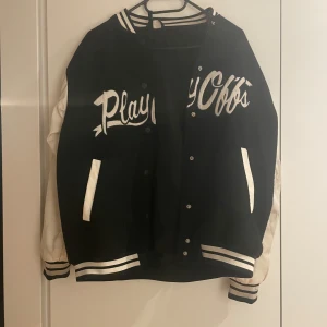 Svart och vit bomberjacka med text - Snygg svart och vit bomberjacka med texten 'Playoffs' på framsidan. Jackan har långa ärmar och ett nummer '92' tryckt på ena ärmen. Den har knappar framtill och ribbade muddar vid ärmslut och nederkant. Den är i fint skick i övrigt men är lite missfärgad på armarna samt har nån fläck, därav det låga priset!