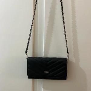 Svart kuvertväska  från Don Donna - Snygg svart axelväska från Don Donna med quiltad design och kedjedetaljer på axelremmen. Väskan har en praktisk dragkedja inuti och är perfekt för att bära dina viktigaste saker. Mått: bredd 21 cm, höjd: 12 cm. 3 invändiga fack varav ett med dragkedja. 