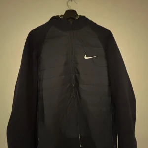 Marinblå jacka från Nike - Snygg svart jacka från Nike med dragkedja och huva. Jackan har en stilren design med ett litet vitt Nike-märke på bröstet. Perfekt för en sportig look.