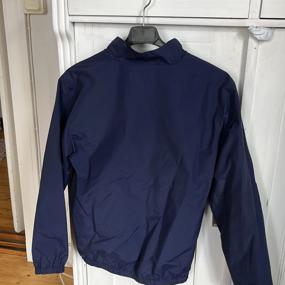 Säljer en stilren mörkblå vindjacka från Lyle & Scott. Jackan har en dragkedja framtill och två fickor med knappar. Den ikoniska loggan syns på bröstet. Perfekt för blåsiga dagar!. Takit.