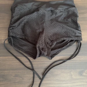 Svarta shorts från SHEIN - Snygga svarta shorts från SHEIN med en unik textur och snörning på sidorna. Perfekta för en trendig look. De har en hög midja och är tillverkade i ett syntetmaterial som ger en bekväm passform.