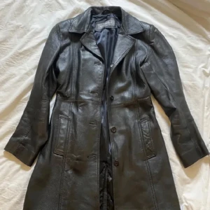 Svart skinnkappa från Roco Baroco - Snygg svart skinnkappa från Roco Baroco med knappar framtill. Kappan har en klassisk krage och långa ärmar, perfekt för en stilren look. Passar bra till både vardag och fest