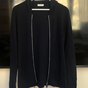 Marinblå hoodie från Sandro - Snygg marinblå hoodie från Sandro med dragkedja framtill. Perfekt för en avslappnad stil. Den har en klassisk design med långa ärmar och en bekväm huva. Defekten på 4e bilden går lätt att laga med bara några stygn eller förslagsvis en lapp. Annars är tröjan i väldigt bra skick, har inte använt den mer än 10 gåmger då den blev för liten. 