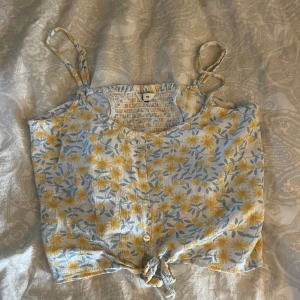 Blommig linnetopp med knyt - Säljer en söt blommig linnetopp i ljusblått och gult med knyt framtill. Toppen har smala axelband och ett elastiskt parti på baksidan för en bekväm passform. Perfekt för varma sommardagar! 🌼