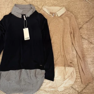 Två tröjor med randig krage från Hailys - Säljer två stilrena tröjor från Hailys. Ens är mörkblå med en randig krage och nederdel i vitt och blått. Den andra är bege med en randig krake och vitt och beiget nertill. Perfekt för en chic och avslappnad look. Tröjan har långa ärmar och en unik design med skjortdetaljer. Båda är i storlek Xs. Den blåa är helt ny med prislapp och den beige är använd en gång. 