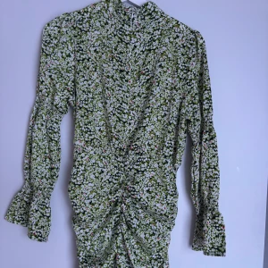 Grön blommig klänning - Säljer en söt grön klänning med blommigt mönster. Klänningen har långa ärmar och en smickrande rynkad detalj framtill. Hör av dig för både frågor och prisförslag!