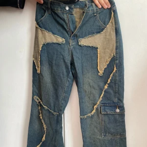 Unika jeans med patchwork-design - Säljer ett par coola jeans med unik patchwork-design och råa kanter. De har en blå bas med kontrasterande beige detaljer och en praktisk ficka på benet. Perfekta för dig som vill sticka ut!