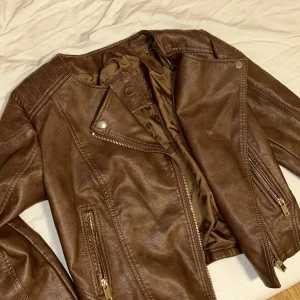 Brun skinnjacka - Snygg brun skinnjacka med dragkedja och stilrena detaljer. Jackan har en klassisk bikerstil med två fickor framtill och en asymmetrisk dragkedja. Perfekt för en cool och avslappnad look.