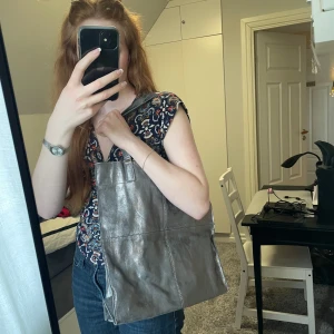 Stor silvrig axelväska  - Snygg och rymlig axelväska i silvrigt glänsande skinn. Perfekt för att bära med sig allt man behöver under dagen, får plats med dator och mycket mer! Lädret har en spricka på ena sidan men annars är den i bra skick🩶