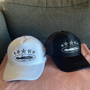 Svart och vit keps med stjärndetaljer - Två kepsar i svart och vitt med broderade stjärnor och ett ovalt emblem framtill. Kepsarna har ett nätmaterial baktill för ventilation och justerbar passform. En keps för 250 och 2 för 400 