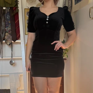Vintage Svart sammetsklänning med knappar - VINTAGE! Köpt secondhand och använd några få gånger. Elegant svart sammet klänning med kort ärm och dekorativa knappar framtill. Klänningen har en figurnära passform och en kort längd, perfekt för festliga tillfällen. 