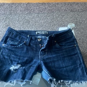 Mörkblå jeansshorts från Original Denim - Snygga mörkblå jeansshorts med slitna detaljer och fransig kant. De har en klassisk femficksdesign och dragkedjor på bakfickorna för en cool look. Perfekta för en avslappnad stil.