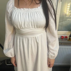 Vit klänning med knytband - Charmig vit klänning med långa ärmar och knytband i midjan. Den har en smickrande passform med rynkad detalj vid bysten och en mjuk, flödande kjol. Perfekt för en somrig look!