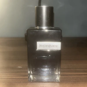 Yves Saint Laurent parfymflaska - Elegant parfymflaska från Yves Saint Laurent i mörk glasdesign med metalliskt lock. Flaskan har en stilren rektangulär form och en tydlig logotyp på framsidan. Perfekt för den som söker en lyxig doftupplevelse. Lite mindre än hälften finns kvar i flaskan. Den är riktig o köpt på ATD Fragrance i Enköping.