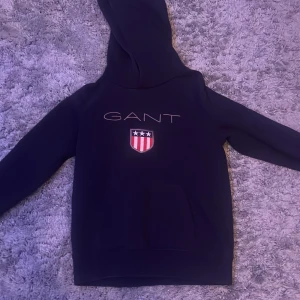 Mörkblå hoodie från GANT - Säljer en snygg mörkblå hoodie från GANT med ett stilrent tryck och ett emblem på bröstet. Tröjan har en klassisk känguruficka och en bekväm huva. Perfekt för en avslappnad stil. Väldigt bra skick då den känns ny