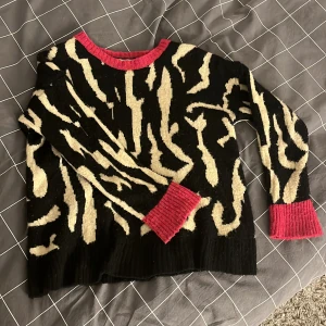 Zebra mönstrad tröja från Isabella D. - Säljer en unik svart tröja med beige mönster och kontrasterande rosa detaljer vid hals och ärmslut från Isabella D. Tillverkad i Italien, perfekt för att sticka ut med stil. Använd ett fåtal gånger men inget som syns. 