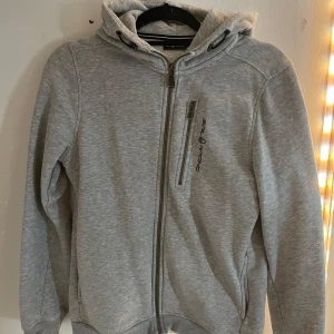 Grå hoodie från Sail Racing - Säljer en stilren grå hoodie från Sail Racing med dragkedja och justerbar huva. Perfekt för kyliga dagar med sin mjuka insida och praktiska fickor. En klassisk och bekväm tröja för alla tillfällen.