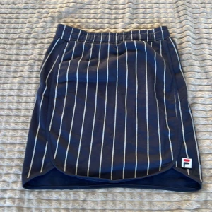 Blå randig kjol från Fila - Snygg blå kjol från Fila med vita ränder. Perfekt för en sportig look. Kjolen har en elastisk midja för extra komfort och en liten slits på sidan. Den är gjord i ett lätt och bekvämt material.