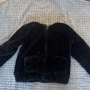 Svart pälsjacka med huva och öron - Mysig svart pälsjacka med huva som har söta öron. Jackan har långa ärmar och stängs med dragkedja. Perfekt för att hålla sig varm och stilren under kyliga dagar.