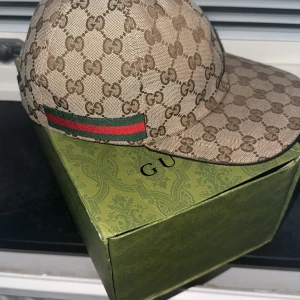 Beige keps från Gucci - Snygg beige keps från Gucci med det ikoniska GG-mönstret. Kepsen har en justerbar rem baktill och en grön och röd rand på sidan. Perfekt för att ge din outfit en lyxig touch.
