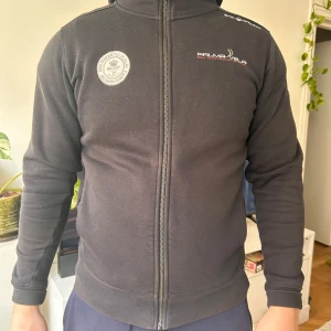 Svart hoodie från Sail Racing - Säljer en svart hoodie från Sail Racing med dragkedja framtill. Tröjan har en hög krage och en liten logotyp på bröstet. Perfekt för en avslappnad stil. Modellen är 185 cm och väger 100 kg. 
