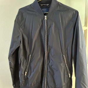 Svart bomberjacka från GANT - Säljer en stilren svart bomberjacka från GANT. Jackan har dragkedja framtill och två fickor med dragkedjor. Perfekt för en cool och avslappnad look.