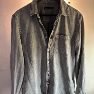 Grå jeansskjorta från Zara - Snygg grå jeansskjorta från Zara med klassisk krage och knappar framtill. Skjortan har en bröstficka och långa ärmar, perfekt för en avslappnad stil. Passar bra till både jeans och chinos.