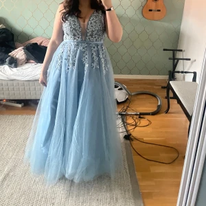 Blå långklänning med spetsdetaljer - Jättefin  blå långklänning med djup v-ringning och vackra spetsdetaljer på livet. Klänningen har en luftig kjol i tyll. Jag säljer pga att jag beställde  klänningen i två storlekar och det var svårt att skicka tillbaka till butiken. Jättefin och bra modell. Perfekt för speciella tillfällen. Dragkedja i ryggen.