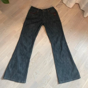Utsvängda jeans från CTM Jean's - Snygga lågmidjade utsvängda jeans i blå denim från CTM Jean's. De har en klassisk femficksdesign med bruna detaljer vid fickorna och en knappgylf. Perfekta för en avslappnad stil.