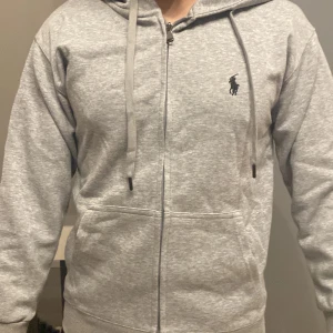Grå hoodie från Polo Ralph Lauren - Säljer en stilren grå hoodie från Polo Ralph Lauren med dragkedja och justerbar huva. Den har en broderad logotyp på bröstet och praktiska fickor framtill. Perfekt för en avslappnad look.