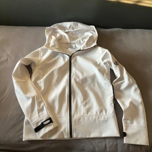 Vit jacka från Moncler - Säljer en stilren vit jacka från Moncler med dragkedja och huva. Jackan har ett Moncler-märke på ärmen och är perfekt för vår och höst. Den har långa ärmar och en modern design.