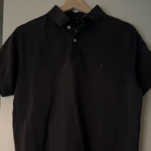 Svart pikétröja från Boomerang - Säljer en klassisk svart pikétröja från Boomerang i storlek M. Tröjan har korta ärmar och en stilren design med knappar vid kragen. Perfekt för en avslappnad och stilren look.