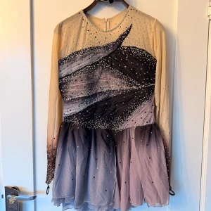 Konståkningsklänning Ny/oanvänd - Säljer en fantastisk konståkningsklänning med glittriga detaljer och pärlor. Klänningen har en ombre-effekt i beige och mörka toner, med långa ärmar och en kort kjol som är längre baktill. Klänningen är helt ny och oanvänd. 
