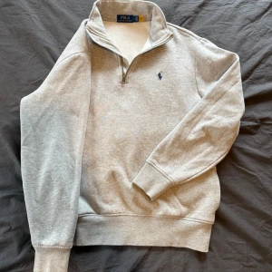 Grå halvzip från Polo Ralph Lauren - Säljer en stilren grå tröja från Polo Ralph Lauren med halv dragkedja och broderad logga på bröstet. Perfekt för en avslappnad look. Tröjan har långa ärmar och är tillverkad i ett mjukt material. Nypris 1900! Använd fåtal gånger.