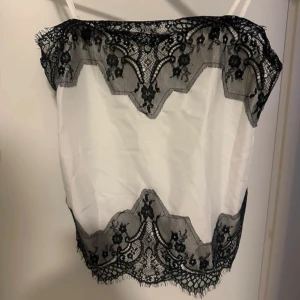 Vit linne med svart spetsdetalj - Gulligt vitt linne med tunna axelband och vacker svart spetsdetalj längs överkanten och nederkanten. Perfekt för en stilren och chic look. Oanvänd storlek S