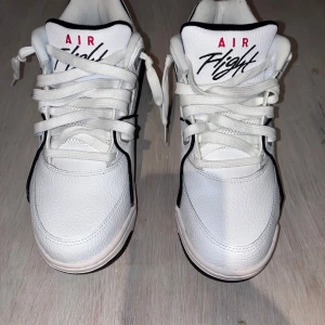 NIKE Air Flight  - Dessa är Nike Air Flight 89 sneakers i vitt med svarta och röda detaljer. De har en hög modell, tydlig “Air Flight”-logga på plösen och en klassisk Nike Swoosh på sidorna. Mellansulan har synlig Air-dämpning, och på baksidan finns “Nike Air”-loggan broderad i rött.
