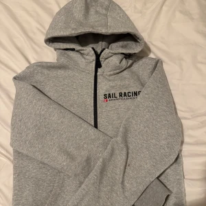 Grå hoodie från Sail Racing - Säljer en grå hoodie från Sail Racing med tryck på bröstet och ryggen. Tröjan har en dragkedja framtill och en stor luva. Perfekt kyligare kvällar/dagar. Storlek S sitter som S. Riktigt skönt material inuti, men säljer då den är för liten. Vid frågor eller funderingar kontakta mig!