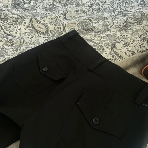 Svarta byxor med knappar - Snygga svarta kostymbyxor med mid waste men skulle ändå kunna säga att dem är som low waste och knappar framtill och där bak. Perfekta för en stilren look. Byxorna har en tight passform och är tillverkade i ett bekvämt material.