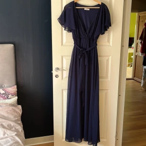 Marinblå långklänning från Goddiva - Elegant marinblå långklänning från Goddiva med v-ringning och volangärmar. Klänningen har ett knytband i midjan för en smickrande passform. Perfekt för festliga tillfällen.