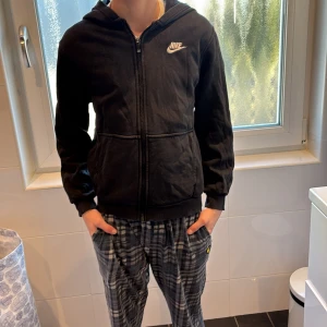 Svart hoodie från Nike - Säljer en svart hoodie från Nike med dragkedja och huva. Perfekt för en avslappnad stil. Den har långa ärmar och en klassisk passform. Nike-loggan är broderad på bröstet. Tröjan har en liten defekt, dragkedjan är smått sliten (se bild 3), men utifrån det har inte tröjan någon annan defekt!