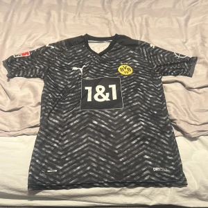 Svart och grå Borussia Dortmund Tröja - Snygg svart och grå fotbollströja från Borussia Dortmund med korta ärmar. Tröjan har ett mönster i grått och svart med gula detaljer och sponsorloggor. Perfekt för fans av laget!