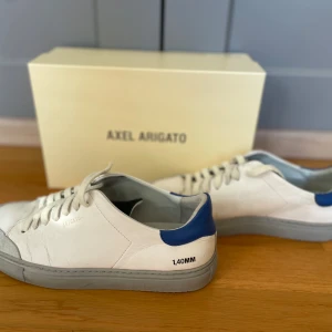 sneakers från Axel Arigato - Snygga vita sneakers från Axel Arigato med blå detaljer och grå sula. Skorna har snörning och en stilren design med märkets logga på sidan. Perfekta för en casual look.