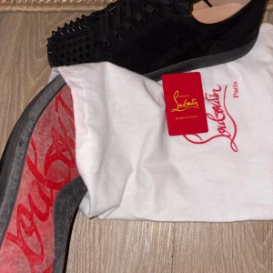 Svarta sneakers med nitar från Christian Louboutin - Snygga svarta sneakers i mocka från Christian Louboutin med ikoniska röda sulor. Skorna har nitar på tån och snörning för en edgy look. 