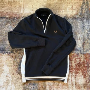 Svart tröja med dragkedja från Fred Perry - Säljer en stilren svart tröja från Fred Perry med dragkedja och vita detaljer längs sidorna och nederkanten. Tröjan har en broderad logga på bröstet och långa ärmar. Perfekt för en casual look! Är i bra skick. Storlek S